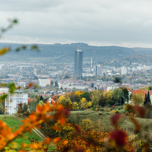 Panoramic view of Jena, Germany, surrounded by wooded hills and autumn foliage. The cityscape features residential and commercial buildings, with the distinctive JenTower rising prominently in the centre beneath an overcast sky.