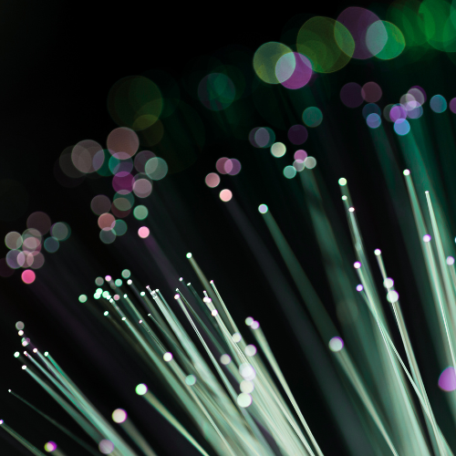 Abstract close-up of illuminated fibre optic strands against a dark background, with glowing points of green, purple, pink and white light. The image represents photonics, optical technology, high-speed data transmission and advanced communications.