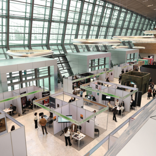 Elevated view of a modern trade show and exhibition hall with multiple company booths, displays and attendees networking throughout the venue. The bright, contemporary space highlights industry events as a place for businesses, specialists and professionals to connect, share ideas and build relationships.