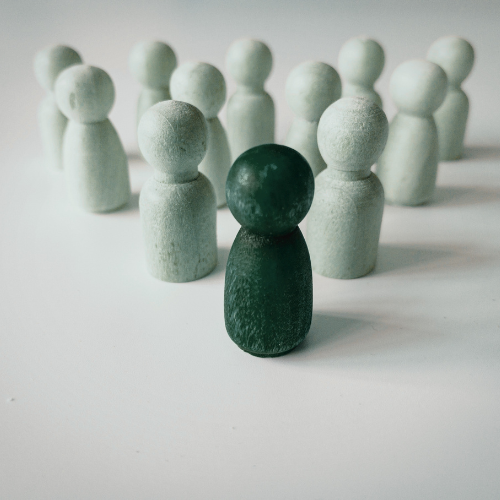 A dark green wooden figure stands prominently in front of a group of lighter figures. The image represents standing out from the crowd, leadership, specialist talent and the importance of finding the right person for a role.